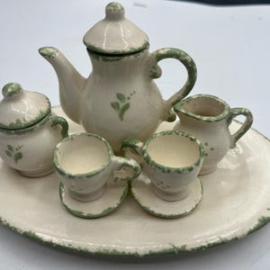 May include: A white ceramic tea set with green accents. The set includes a teapot, creamer, sugar bowl, and two teacups and saucers. The set is on a white oval tray with a green rim.