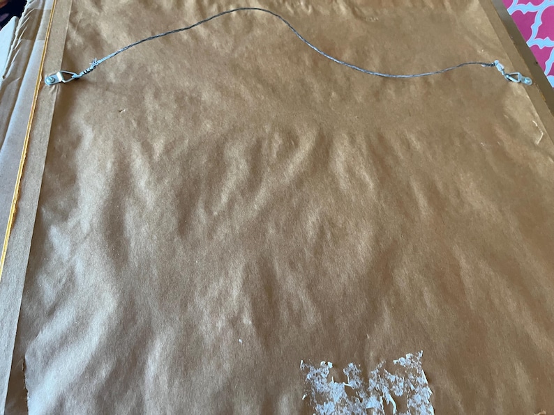 May include: The reverse of a framed picture, with a brown paper backing. A thin, silver wire is stretched across the top for hanging. Metal clips secure the wire to the frame. The paper has some white adhesive residue.