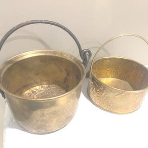 May include: Two antique brass pots with handles. The larger pot has a dark metal handle and a rounded shape. The smaller pot has a decorative hammered design and a curved handle. Both pots are on a white surface.