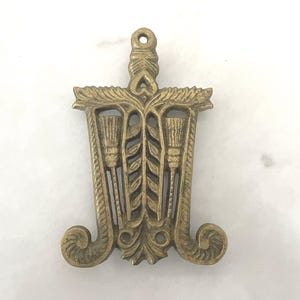 May include: A brass trivet featuring an intricate openwork design with a central vertical leaf motif flanked by two stylized broom icons. The piece has a rectangular frame with decorative scrollwork at the base and a small circular hanging hole at the top.