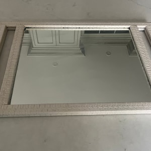 May include: A white rectangular mirror with a textured frame. The mirror is reflecting a white ceiling with recessed lighting.