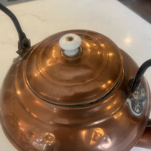 May include: Close-up of a copper tea kettle with a white ceramic knob on the lid. The kettle has a black handle and spout. The copper surface reflects light, highlighting its curves and details.