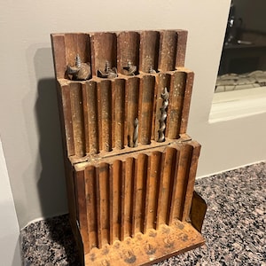 May include: A wooden drill bit holder with multiple slots for storing drill bits. The holder is made of dark wood and has a worn, rustic appearance.