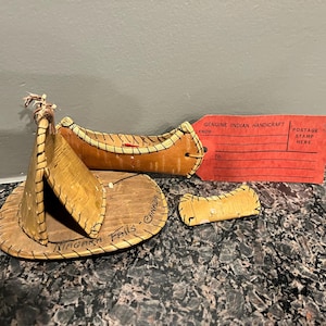 May include: A miniature wooden canoe with a red stripe and the words "Niagara Falls - Canada" written on it. The canoe is part of a set of three pieces, including a small wooden paddle and a miniature wooden tent. A red tag with the words "Genuine Indian Handicraft" is attached to the canoe.