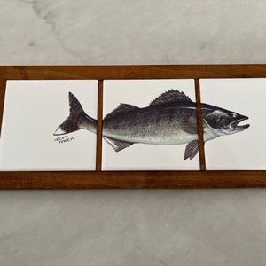 May include: A wooden trivet with three white ceramic tiles. The center tile features a detailed illustration of a fish with the artist's signature "Geoff Walker" in the bottom left corner.