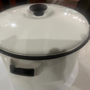 May include: A white enamel pot with a black rim and handles. The pot has a matching lid with a black knob. This kitchenware item is suitable for cooking and food storage.