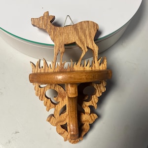 May include: Wooden deer shelf with a small round shelf and decorative carved details. The deer is standing on a grassy ledge. The shelf is made of light brown wood and has a decorative bracket.