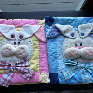 May include: Two decorative Easter bunny pillows. One pillow has a pink and yellow patchwork design, with a bunny face featuring button eyes and a plaid bow. The other pillow has a blue denim patchwork design, with a bunny face and a blue plaid bow.
