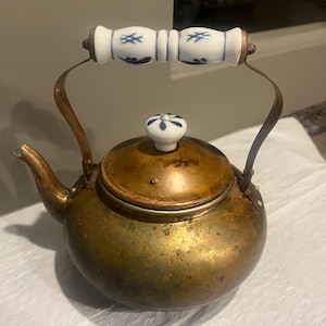 May include: Antique brass teapot with a white and blue porcelain handle and lid knob. The teapot has a curved spout and a rounded body. The handle is made of brass and curves upwards. The lid knob is a small, round porcelain piece with a blue floral design.