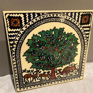May include: Square, hand-painted ceramic tile featuring a stylized tree with green leaves and red fruit. The tree is surrounded by deer and a boar. The border has geometric and floral patterns in black, blue, and orange.