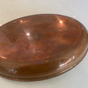 May include: An oval-shaped copper roasting pan with a polished, reflective metallic surface. The pan features a smooth, rounded rim and a dark, curved handle extending from one side, showcasing a warm, reddish-brown metallic finish throughout.