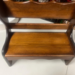 May include: A small, wooden step stool with a dark brown finish. The stool has a curved back and two horizontal supports. The seat is rectangular and the legs are short and sturdy. The wood grain is visible.