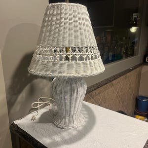 May include: White wicker table lamp with a woven shade and base. The lamp has a classic design with a flared base and a tapered shade. The lamp is plugged into an electrical outlet. The lamp is approximately 24 inches tall.