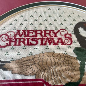 May include: Cross-stitch artwork with a Christmas theme. The words "MERRY CHRISTMAS" are stitched in red. A goose with brown and black feathers and green accents is also featured in the design. The artwork is framed.
