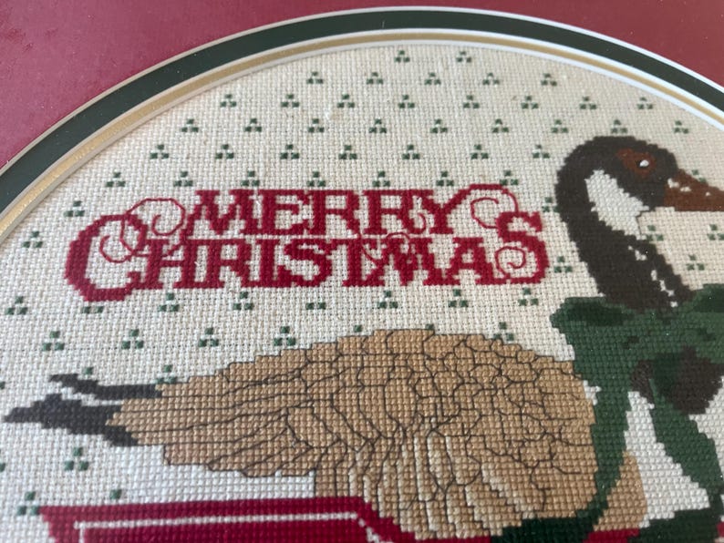 May include: Cross-stitch artwork with a Christmas theme. The words "MERRY CHRISTMAS" are stitched in red. A goose with brown and black feathers and green accents is also featured in the design. The artwork is framed.