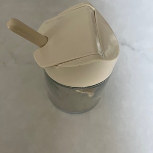 May include: A white plastic sugar dispenser with a clear glass container and a small white plastic spoon.