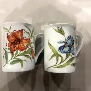 May include: Two white ceramic mugs with floral designs. One mug features a red and orange lily, while the other mug features a blue iris.