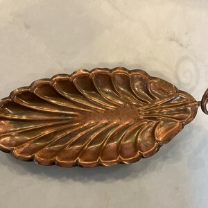 May include: A copper serving dish with a scalloped edge and a decorative handle. The dish is oval shaped and has a shiny finish.