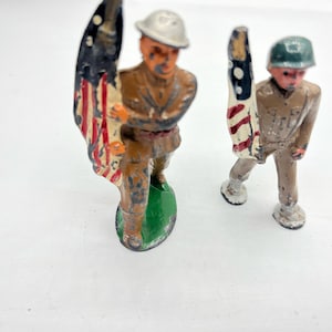 May include: Two vintage toy soldiers holding American flags. The soldier on the left is wearing a helmet and is holding a flag with red, white, and blue stripes. The soldier on the right is wearing a helmet and is holding a flag with red, white, and blue stars. Both soldiers are made of metal and are painted in a realistic style.