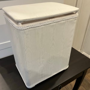 May include: White rectangular laundry hamper with a fabric cover and a rope trim around the top edge. The hamper is sitting on a dark brown table.