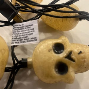 May include: A string of decorative lights featuring multiple off-white, textured skull-shaped covers connected by a black electrical cord. A white rectangular safety information tag is attached to the cord, detailing electrical specifications.