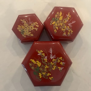 May include: Three red hexagonal boxes with floral designs. The boxes are nested inside each other, with the largest box on the bottom and the smallest box on top. The boxes are decorated with yellow flowers and green leaves on a red background.