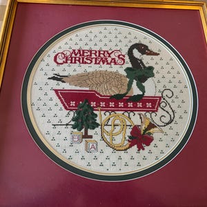 May include: Framed cross-stitch artwork in a gold frame. The artwork shows a goose in a sleigh with the words "MERRY CHRISTMAS". The goose wears a green bow. A Christmas tree and a horn are also depicted.
