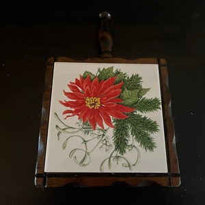 May include: A wooden hot pad with a ceramic tile featuring a red poinsettia flower and green pine branches on a white background.
