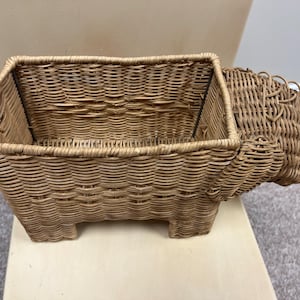 May include: A woven wicker basket shaped like a dog. The basket is light brown and has a rectangular opening. The basket is sitting on a light brown surface.