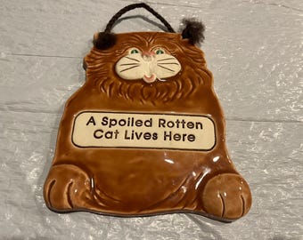 May include: Brown ceramic cat-shaped wall plaque with the text "A Spoiled Rotten Cat Lives Here". The cat has a white face, green eyes, and black whiskers. It has a black string for hanging.