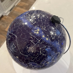 May include: A spherical puzzle depicting a starry night sky with constellations. The puzzle is primarily dark blue with white and yellow star patterns and constellation lines. A black power cord and plug are attached to the top of the puzzle.
