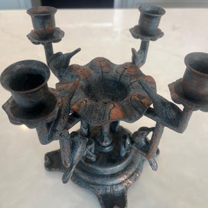 May include: A dark, weathered metal candelabra featuring four candle holders supported by bird-shaped figures. The central structure has a scalloped, flower-like design with a distressed finish showing hints of copper and bronze tones throughout.