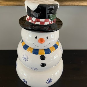 May include: A white ceramic snowman cookie jar with a black top hat, blue and yellow scarf, and blue snowflake details.