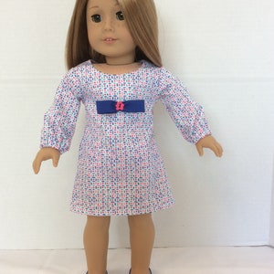 May include: A doll wearing a white dress with a blue and pink geometric pattern. The dress has a blue bow with a pink flower button. The doll is wearing blue shoes with white bows.