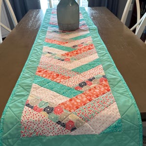 May include: A turquoise and coral quilted table runner with a chevron pattern. The runner is made from various patterned fabrics, including floral, polka dot, and geometric prints.