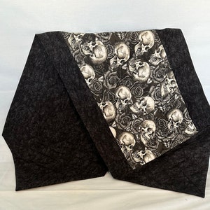 May include: Black and white table runner with a skull and rose design. The runner has a black background with a white skull and rose pattern. The runner is made of fabric and has a fringed edge.