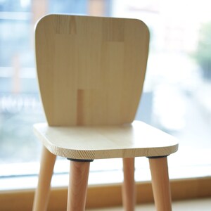 May include: A light-colored wooden chair featuring a smooth, flat seat and a rounded, upright backrest. The chair is supported by four tapered, cylindrical legs that are attached to the underside of the seat with dark circular connectors.