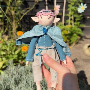 Könnte beinhalten: Eine handgefertigte Puppe mit rosa Haaren, spitzen Ohren und rosigen Wangen, bekleidet mit einem blauen Langarmshirt, einem hellblauen Umhang mit blauem Edelsteinverschluss und beigefarbener Hose. Die Puppe trägt ein kleines Schwert am Hosenbund.