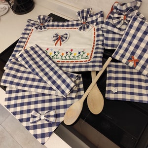 Op de afbeelding: Een set blauwe en witte gingham keukentextiel, inclusief een pannenlappen, een theedoek en een placemat. De textiel is versierd met geborduurde bloemen en strikken. Er zijn ook twee houten lepels.