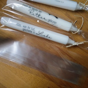 May include: Three white candles wrapped in clear plastic bags with twine. The candles are decorated with black text that reads "Euch ist heute der Retter geboren" and "Alles, was ihr tut, geschehe in Liebe".