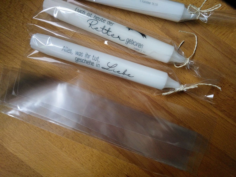 May include: Three white candles wrapped in clear plastic bags with twine. The candles are decorated with black text that reads "Euch ist heute der Retter geboren" and "Alles, was ihr tut, geschehe in Liebe".