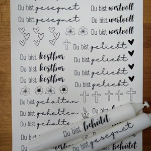 May include: A white sheet of paper with black ink printed on it. The text reads "Du bist gesegnet" (You are blessed) and "Du bist wertvoll" (You are valuable). There are also images of hearts, flowers, and crosses. Three white candles are on the paper, with black ink printed on them. The text on the candles reads "Du bist beh&uuml;tet" (You are protected) and "Du bist gesegnet" (You are blessed).