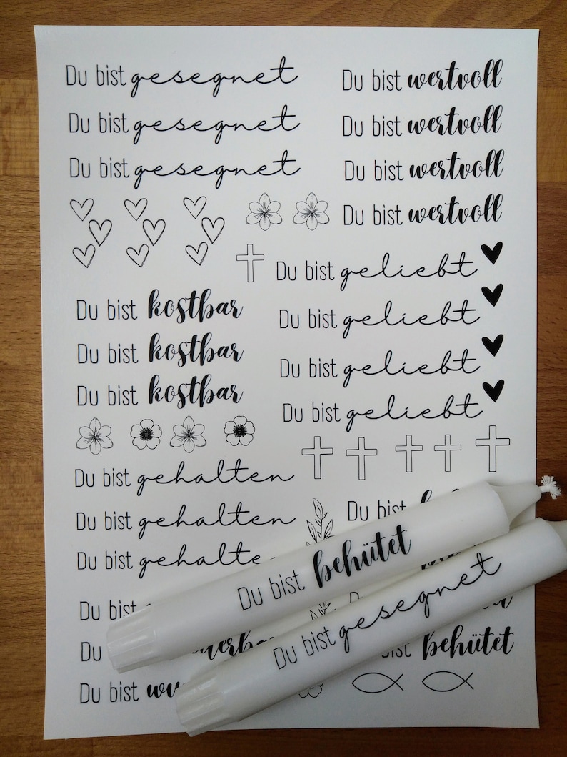 May include: A white sheet of paper with black ink printed on it. The text reads "Du bist gesegnet" (You are blessed) and "Du bist wertvoll" (You are valuable). There are also images of hearts, flowers, and crosses. Three white candles are on the paper, with black ink printed on them. The text on the candles reads "Du bist beh&uuml;tet" (You are protected) and "Du bist gesegnet" (You are blessed).