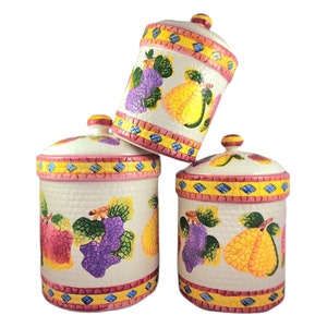 Peut inclure: Ensemble de trois pots en céramique avec couvercles, ornés d'un motif de fruits colorés. Les pots sont blancs avec un motif en mosaïque rose, jaune et bleu autour du haut et du bas. Le motif de fruits comprend des poires, des raisins et des fraises.