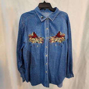 May include: A long-sleeved denim shirt with a button-down front. The shirt features embroidered red cardinals perched on autumn leaves. The collar also has a small embroidered design. The shirt is hanging on a black hanger.
