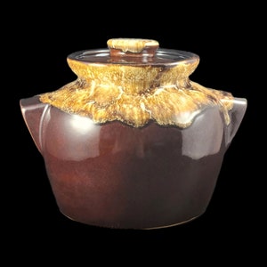 May include: A brown glazed ceramic casserole dish with a lid. The dish has a yellow and brown glaze pattern. The dish has two handles on the sides.