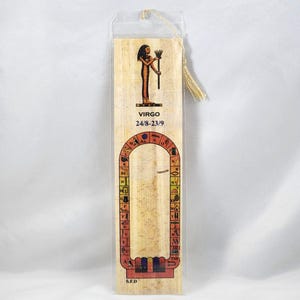 May include: A papyrus bookmark featuring an Egyptian Virgo design. The bookmark has a gold tassel and is encased in clear plastic. The design includes an illustration of a figure, hieroglyphs, and the text "VIRGO 24/8-23/9".