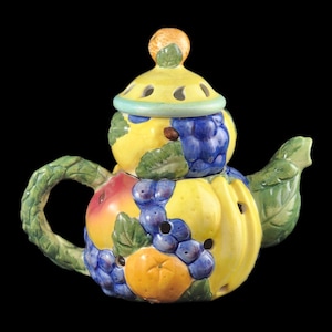 Op de afbeelding: Een keramische theepot versierd met een kleurrijk fruitontwerp. De theepot heeft een geel lichaam met een groene handgreep en tuit. Het fruitontwerp omvat citroenen, sinaasappels, druiven en appels.