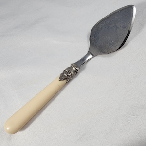 May include: A vintage pie server with a stainless steel blade and a decorative handle. The handle features a cream-colored, rounded grip and a silver-toned, ornate design at the base of the blade. The server is designed for serving pies and cakes.