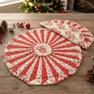 May include: Two quilted Christmas placemats with a red and white poinsettia design. The round placemat has red stripes radiating from the center. The second placemat is a half-circle shape. The placemats are on a wooden table with Christmas decorations in the background.